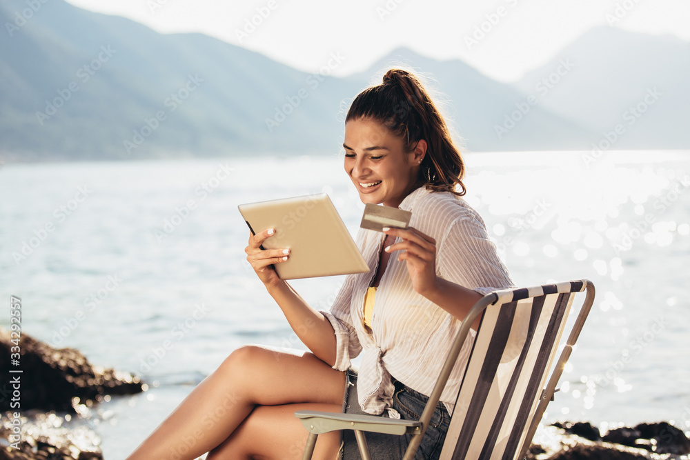 Smiling woman sitting on deck chair by the sea using tablet and credit card on a sunny day