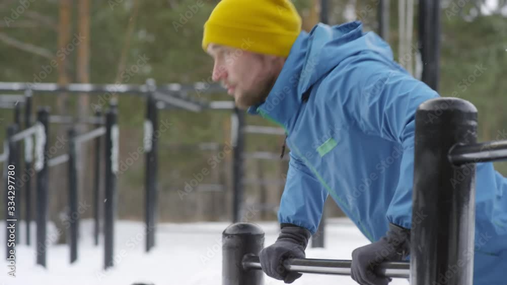 Medium shot of active man in blue windbreaker and yellow beanie hat doing push-ups on workout bar at outdoor gym in winter Medium shot of active man in blue windbreaker and yellow beanie hat doing push-ups on workout bar at outdoor gym in winter