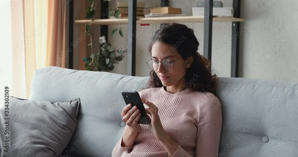 Relaxed young woman using smart phone surfing social media, checking news, playing mobile games or texting messages sitting on sofa. Millennial lady spending time at home with cell gadget technology.