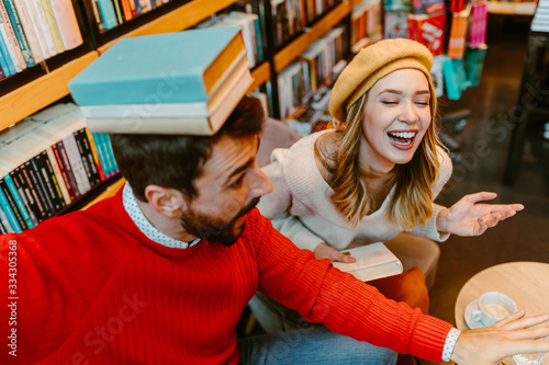 Young student couple is having fun with books in the library. Man is holding some books on his head.