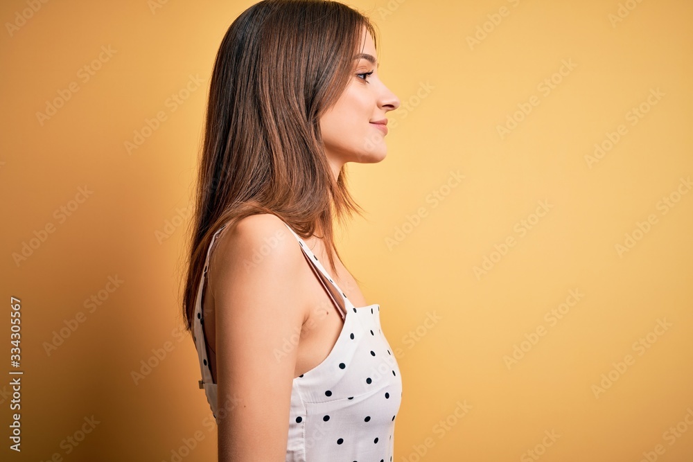 Young beautiful brunette woman wearing casual dress standing over yellow background looking to side, relax profile pose with natural face with confident smile. Young beautiful brunette woman wearing casual dress standing over yellow background looking to side, relax profile pose with natural face with confident smile.