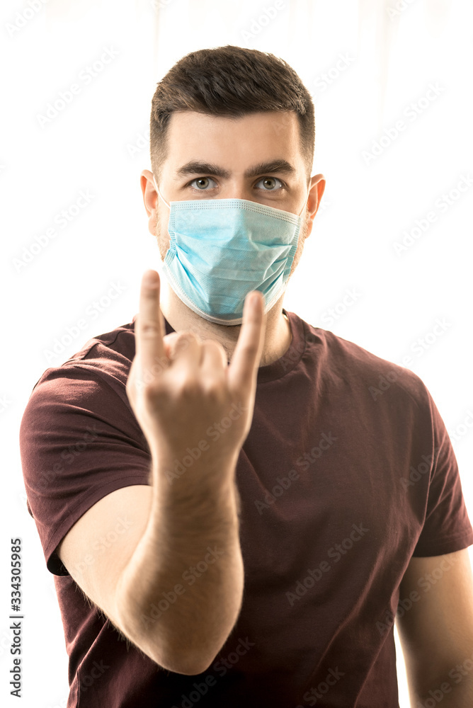 The topic of coronavirus and safety. A young man in a mask from a virus shows a goat sign, on a light background. Vertical frame The topic of coronavirus and safety. A young man in a mask from a virus shows a goat sign, on a light background. Vertical frame