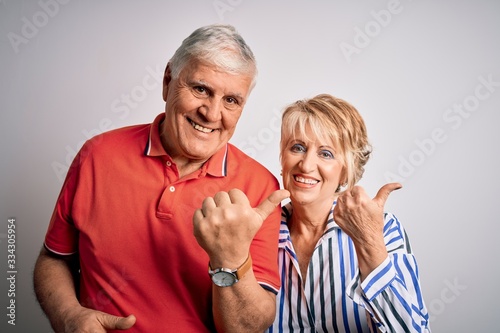 Senior beautiful couple standing together over isolated white background Pointing to the back behind with hand and thumbs up, smiling confident