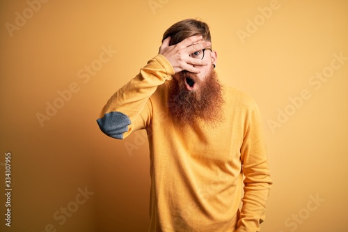 Handsome Irish redhead man with beard wearing glasses over yellow isolated background peeking in shock covering face and eyes with hand, looking through fingers with embarrassed expression.