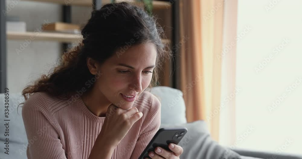 Smiling millennial woman watching funny video stories on smart phone at home. Happy female customer doing mobile shopping, reading news, using apps, surfing internet looking at cell sitting on couch.