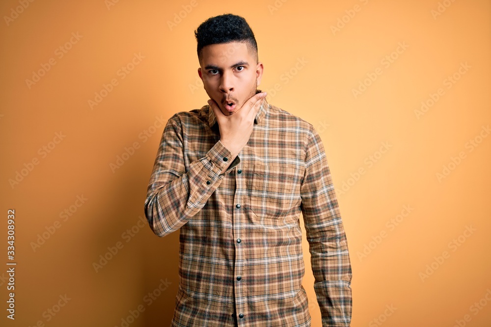 Young handsome man wearing casual shirt standing over isolated yellow background Looking fascinated with disbelief, surprise and amazed expression with hands on chin Young handsome man wearing casual shirt standing over isolated yellow background Looking fascinated with disbelief, surprise and amazed expression with hands on chin