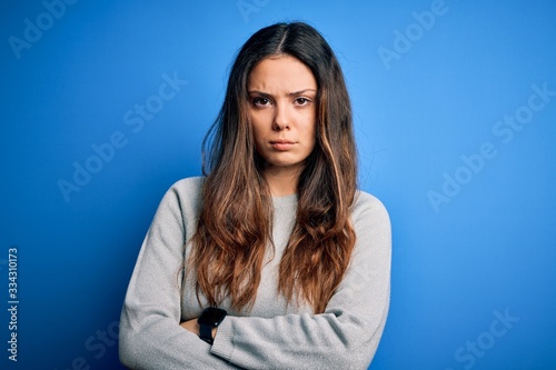 Young beautiful brunette woman wearing casual sweater standing over blue background skeptic and nervous, disapproving expression on face with crossed arms. Negative person.
