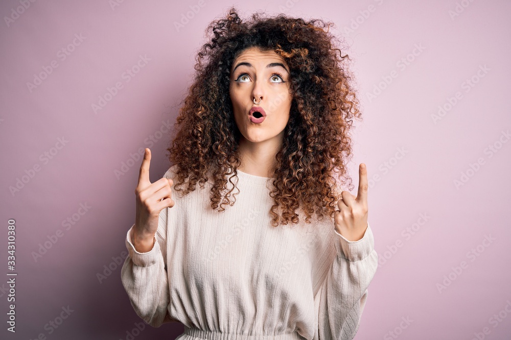 Beautiful woman with curly hair and piercing wearing casual sweater over pink background amazed and surprised looking up and pointing with fingers and raised arms. Beautiful woman with curly hair and piercing wearing casual sweater over pink background amazed and surprised looking up and pointing with fingers and raised arms.