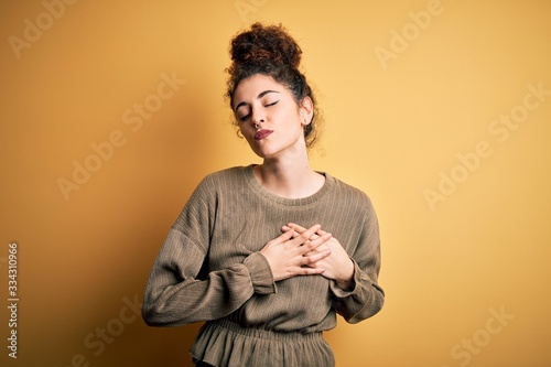 Young beautiful brunette woman with curly hair and piercing wearing casual dress smiling with hands on chest with closed eyes and grateful gesture on face. Health concept. © Krakenimages.com