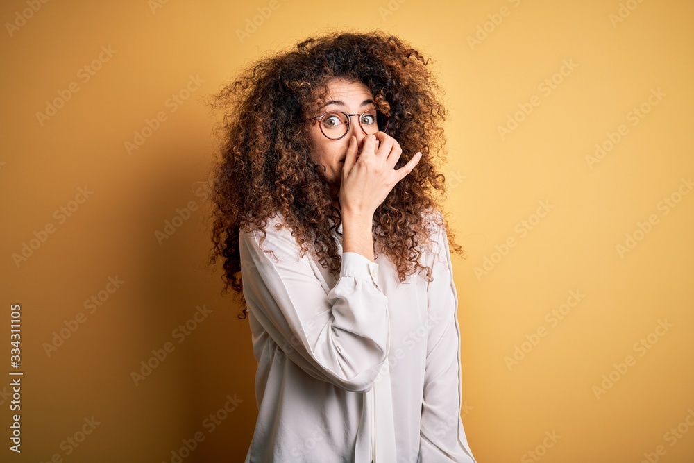 Young beautiful brunette woman with curly hair and piercing wearing shirt and glasses smelling something stinky and disgusting, intolerable smell, holding breath with fingers on nose. Bad smell Young beautiful brunette woman with curly hair and piercing wearing shirt and glasses smelling something stinky and disgusting, intolerable smell, holding breath with fingers on nose. Bad smell