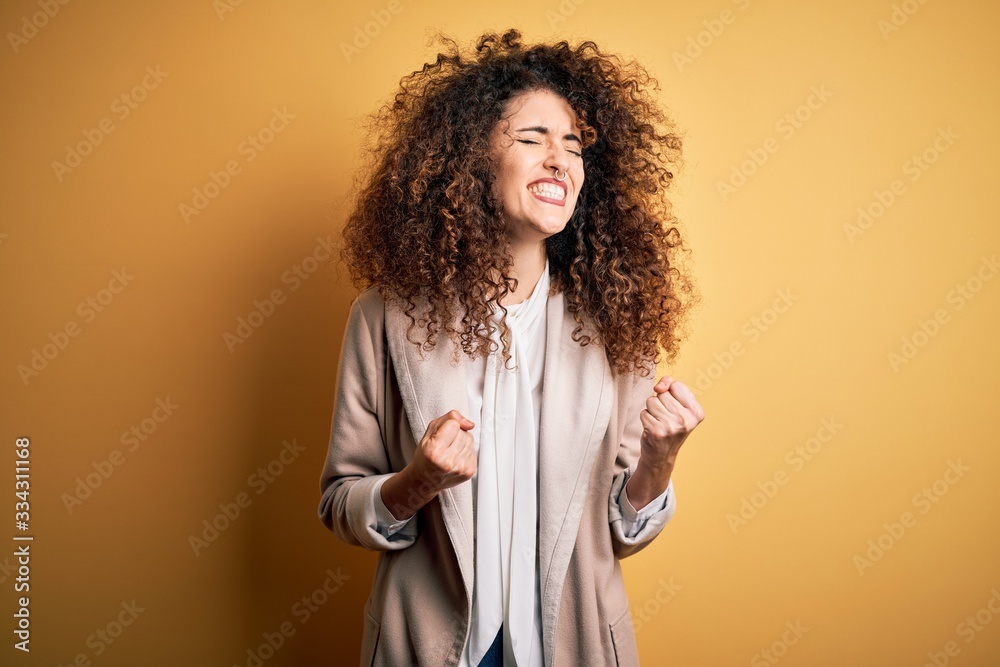 Young beautiful brunette woman with curly hair and piercing wearing casual t-shirt and diadem excited for success with arms raised and eyes closed celebrating victory smiling. Winner concept. Young beautiful brunette woman with curly hair and piercing wearing casual t-shirt and diadem excited for success with arms raised and eyes closed celebrating victory smiling. Winner concept.