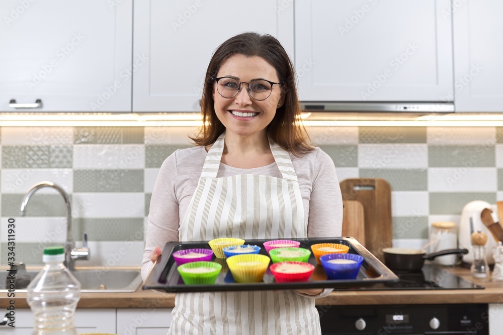 Mature woman in apron with baking tray with cupcakes being prepared Mature woman in apron with baking tray with cupcakes being prepared