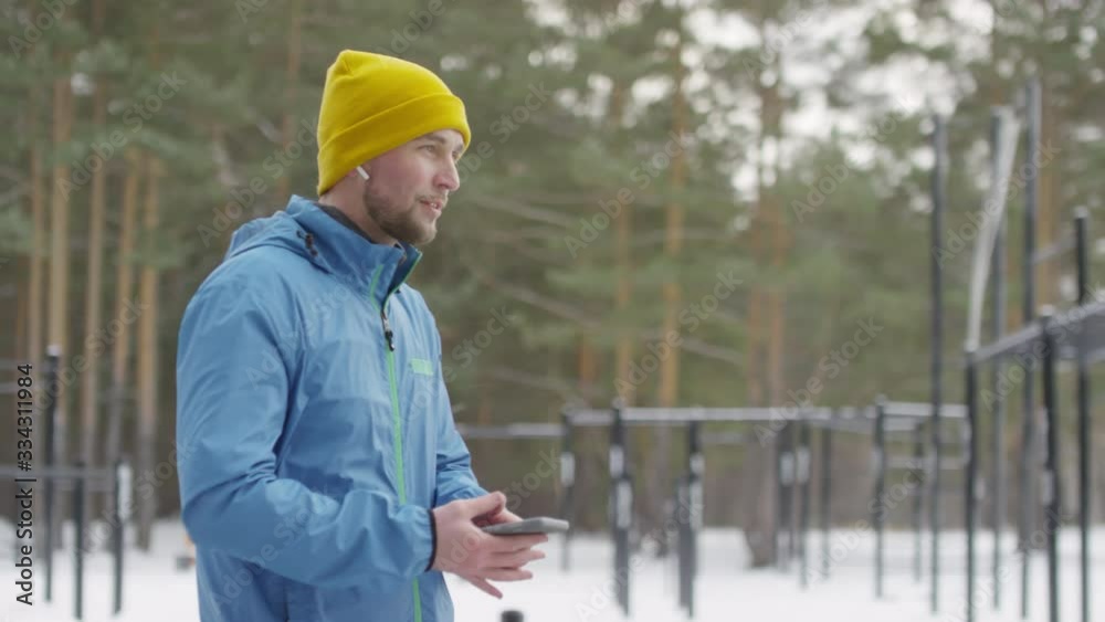 Medium shot of handsome man in wireless earphones choosing song on mobile phone, then smiling and starting his jog outdoors on windy winter day Medium shot of handsome man in wireless earphones choosing song on mobile phone, then smiling and starting his jog outdoors on windy winter day