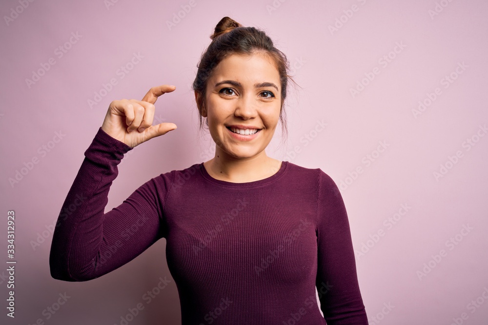 Fototapeta premium Beautiful young woman wearing casual bun hairstyle over pink isolated background smiling and confident gesturing with hand doing small size sign with fingers looking and the camera. Measure concept.