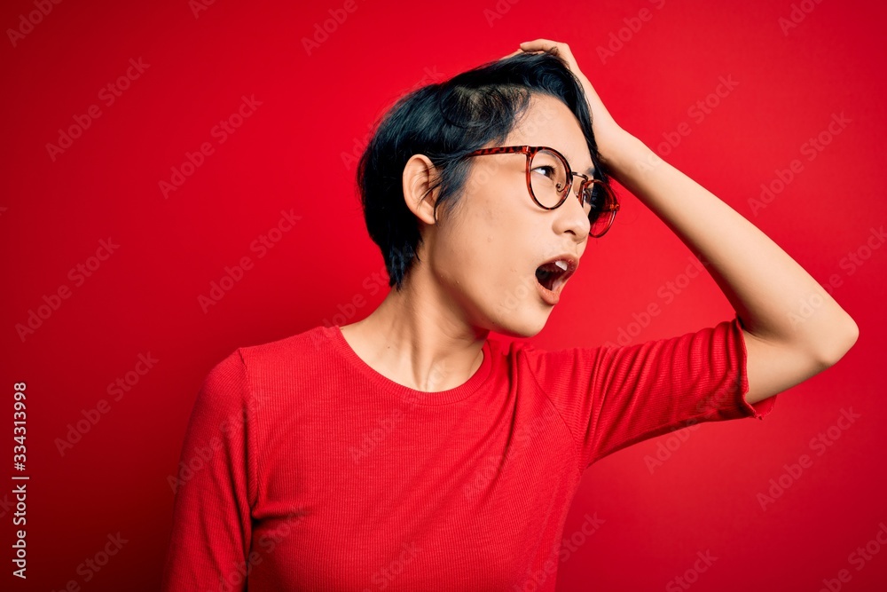 Young beautiful asian girl wearing casual t-shirt and glasses over isolated red background surprised with hand on head for mistake, remember error. Forgot, bad memory concept. Young beautiful asian girl wearing casual t-shirt and glasses over isolated red background surprised with hand on head for mistake, remember error. Forgot, bad memory concept.
