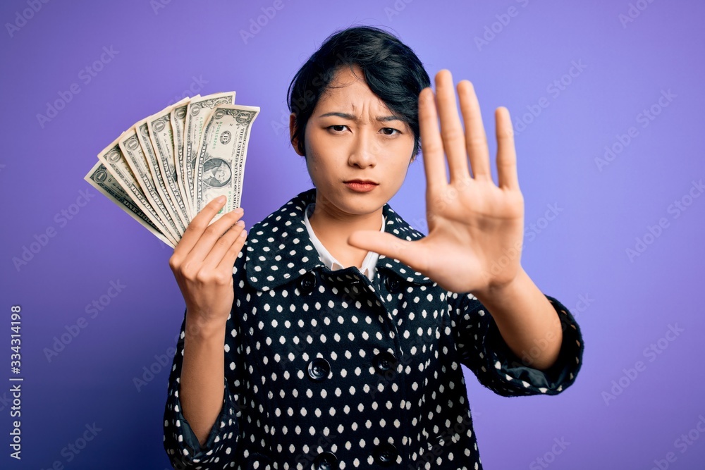 Young beautiful asian girl holding bunch of dollars banknotes over purple background with open hand doing stop sign with serious and confident expression, defense gesture Young beautiful asian girl holding bunch of dollars banknotes over purple background with open hand doing stop sign with serious and confident expression, defense gesture