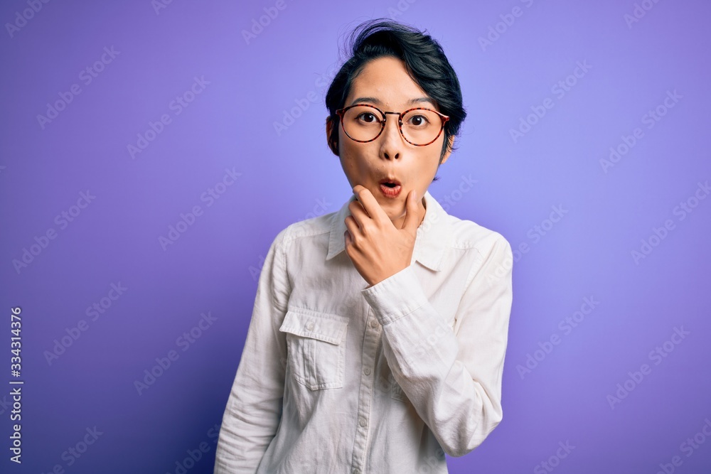 Young beautiful asian girl wearing casual shirt and glasses standing over purple background Looking fascinated with disbelief, surprise and amazed expression with hands on chin Young beautiful asian girl wearing casual shirt and glasses standing over purple background Looking fascinated with disbelief, surprise and amazed expression with hands on chin