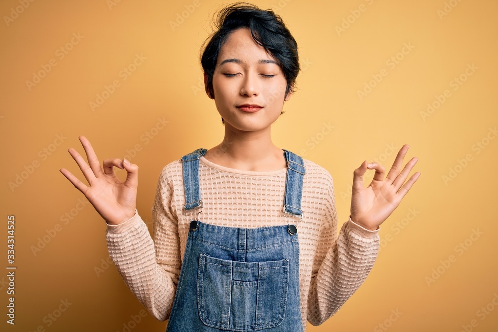 Young beautiful asian girl wearing casual denim overalls over isolated yellow background relax and smiling with eyes closed doing meditation gesture with fingers. Yoga concept. Young beautiful asian girl wearing casual denim overalls over isolated yellow background relax and smiling with eyes closed doing meditation gesture with fingers. Yoga concept.