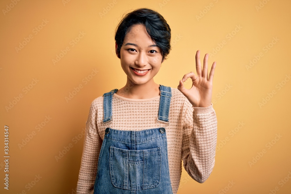 Young beautiful asian girl wearing casual denim overalls over isolated yellow background smiling positive doing ok sign with hand and fingers. Successful expression. Young beautiful asian girl wearing casual denim overalls over isolated yellow background smiling positive doing ok sign with hand and fingers. Successful expression.