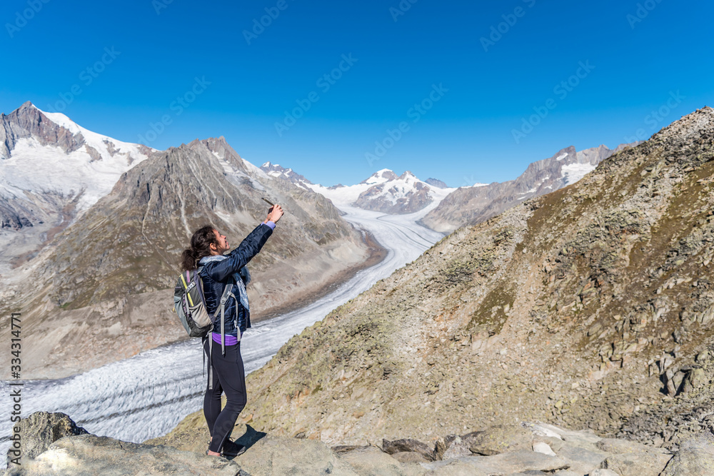 Young Caucasian woman with a backpack on her back, taking a selfie with the monumental Aletsch Glacier behind her. Young Caucasian woman with a backpack on her back, taking a selfie with the monumental Aletsch Glacier behind her.