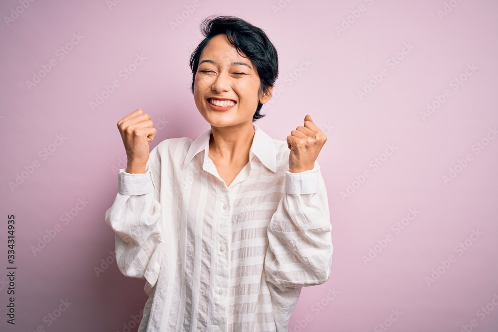 Young beautiful asian girl wearing casual shirt standing over isolated pink background excited for success with arms raised and eyes closed celebrating victory smiling. Winner concept. Young beautiful asian girl wearing casual shirt standing over isolated pink background excited for success with arms raised and eyes closed celebrating victory smiling. Winner concept.
