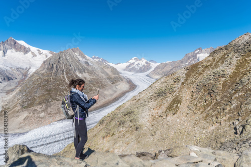 Young Caucasian woman with a backpack on her back, taking a selfie with the monumental Aletsch Glacier behind her.