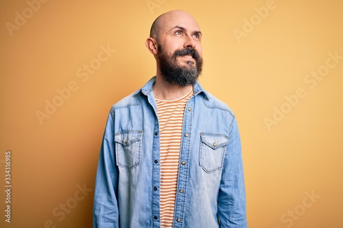 Handsome bald man with beard wearing casual denim jacket and striped t-shirt looking away to side with smile on face, natural expression. Laughing confident.