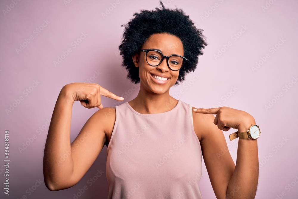 Young beautiful African American afro woman with curly hair wearing t-shirt and glasses looking confident with smile on face, pointing oneself with fingers proud and happy.