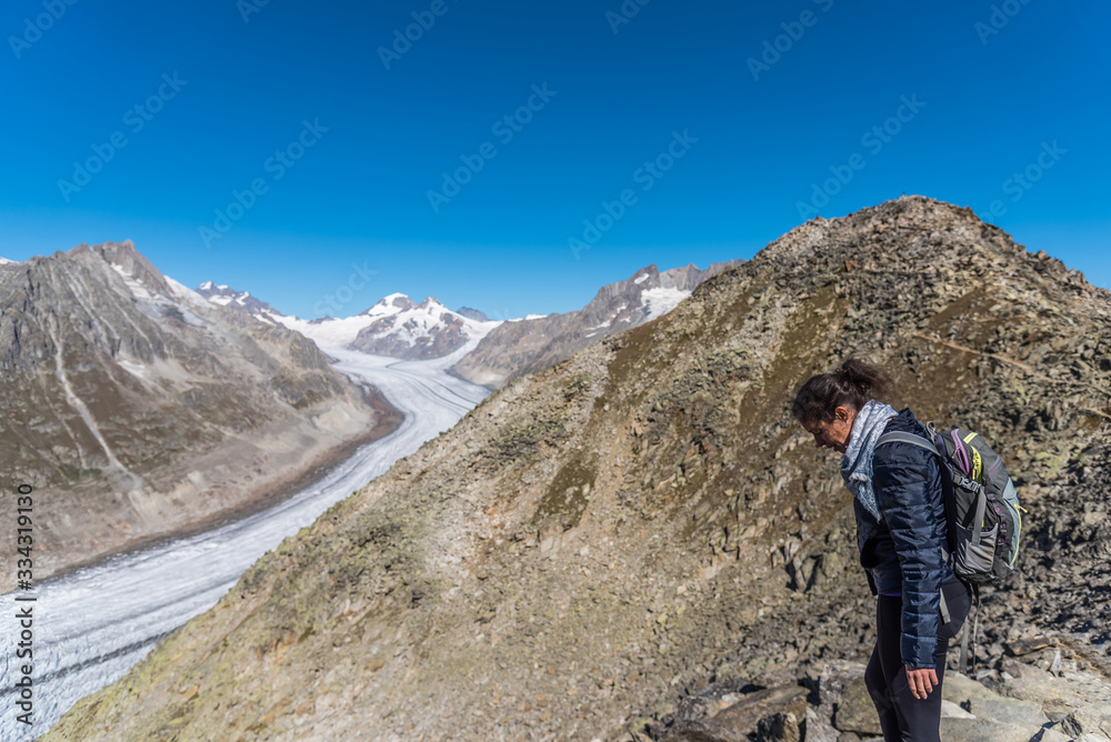 Young Caucasian woman with a backpack on her back looking down at the monumental Aletsch Glacier.This giant stream of ice, which stretches over 23 km