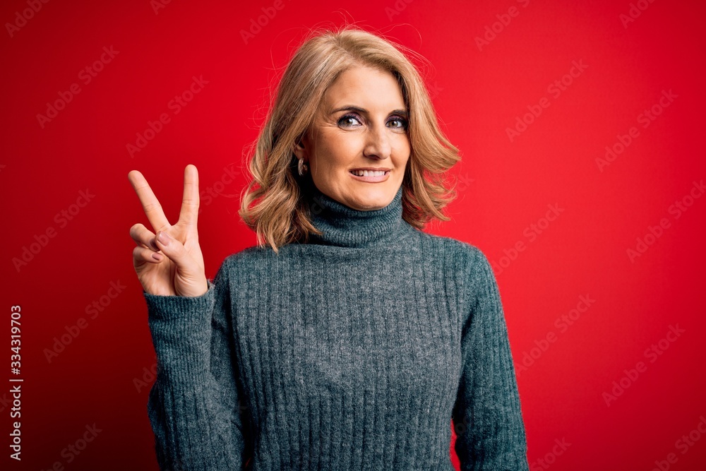 Middle age beautiful blonde woman wearing casual turtleneck sweater over red background smiling with happy face winking at the camera doing victory sign. Number two. Middle age beautiful blonde woman wearing casual turtleneck sweater over red background smiling with happy face winking at the camera doing victory sign. Number two.