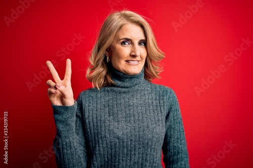 Middle age beautiful blonde woman wearing casual turtleneck sweater over red background smiling with happy face winking at the camera doing victory sign. Number two. © Krakenimages.com