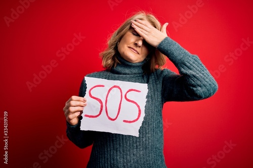 Middle age beautiful blonde woman holding paper with sos message over red bakground stressed with hand on head, shocked with shame and surprise face, angry and frustrated. Fear and upset for mistake.