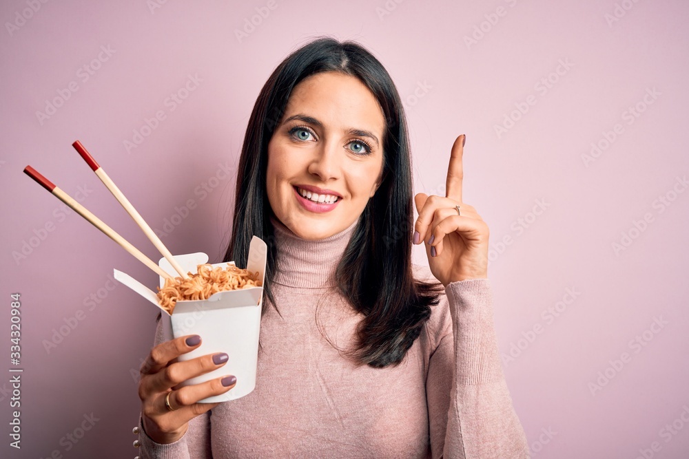 Young brunette woman holding delivery takeaway paper box over yellow background surprised with an idea or question pointing finger with happy face, number one Young brunette woman holding delivery takeaway paper box over yellow background surprised with an idea or question pointing finger with happy face, number one