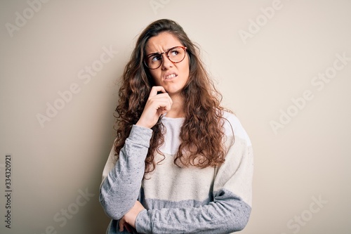 Young beautiful woman with curly hair wearing sweater and glasses over white background with hand on chin thinking about question, pensive expression. Smiling with thoughtful face. Doubt concept.