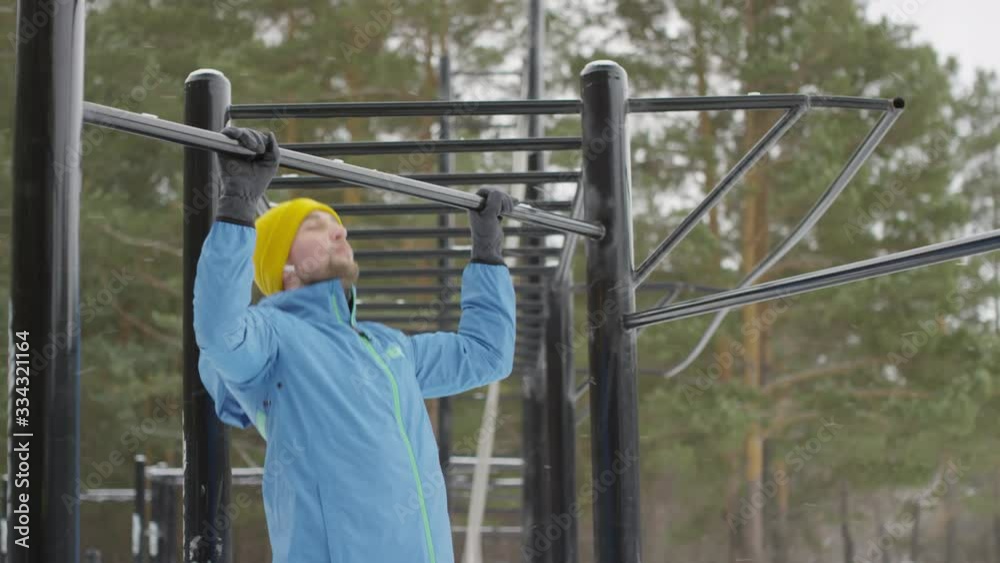 Handheld tilt up shot of sporty man in blue windbreaker and yellow hat doing pull-ups on workout bar at outdoor gym in winter Handheld tilt up shot of sporty man in blue windbreaker and yellow hat doing pull-ups on workout bar at outdoor gym in winter
