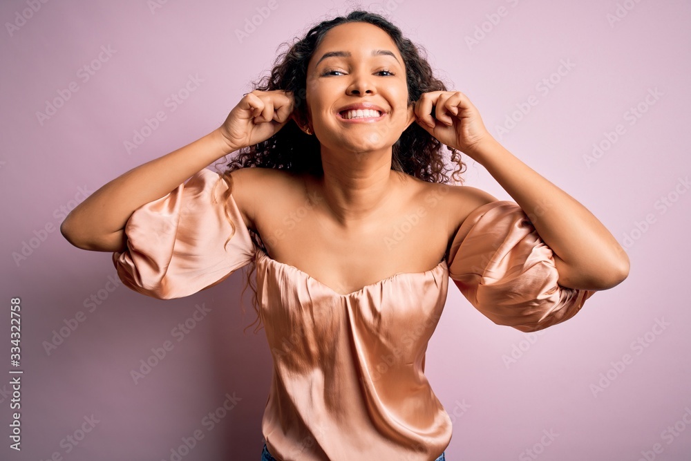 Fototapeta premium Young beautiful woman with curly hair wearing casual t-shirt standing over pink background Smiling pulling ears with fingers, funny gesture. Audition problem