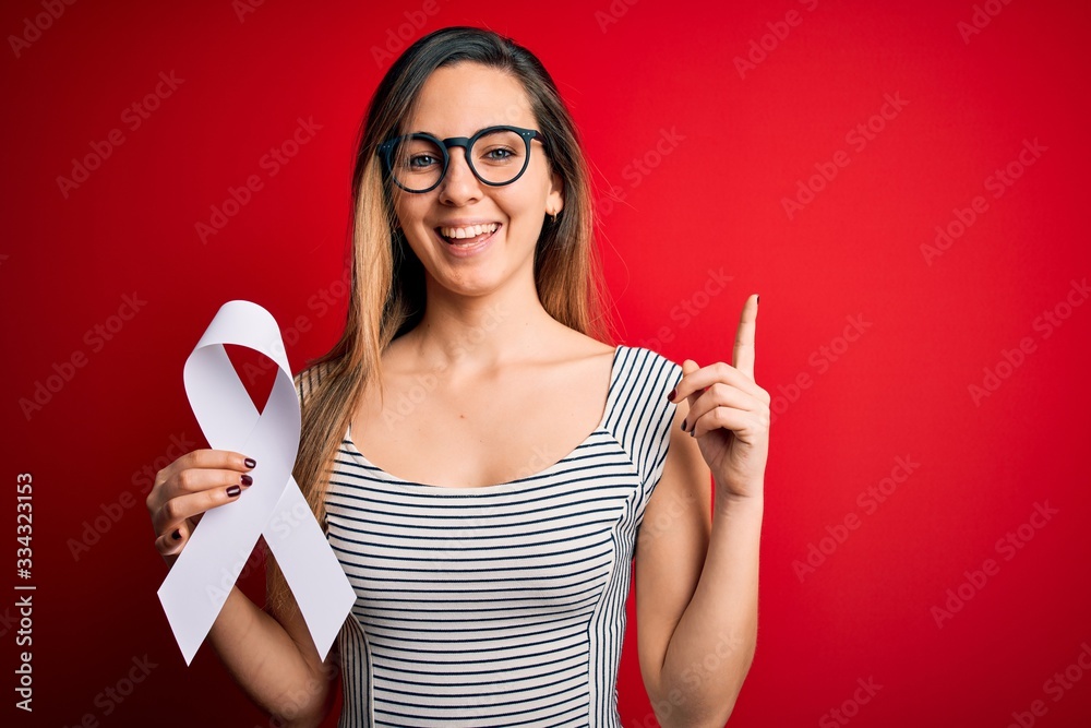 Young beautiful blonde woman with blue eyes holding white ribbon over red background surprised with an idea or question pointing finger with happy face, number one Young beautiful blonde woman with blue eyes holding white ribbon over red background surprised with an idea or question pointing finger with happy face, number one