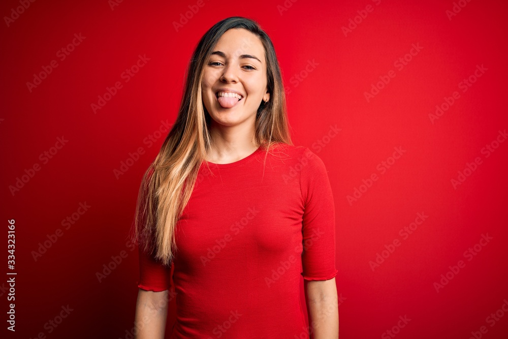 Young beautiful blonde woman with blue eyes wearing casual t-shirt over red background sticking tongue out happy with funny expression. Emotion concept. Young beautiful blonde woman with blue eyes wearing casual t-shirt over red background sticking tongue out happy with funny expression. Emotion concept.