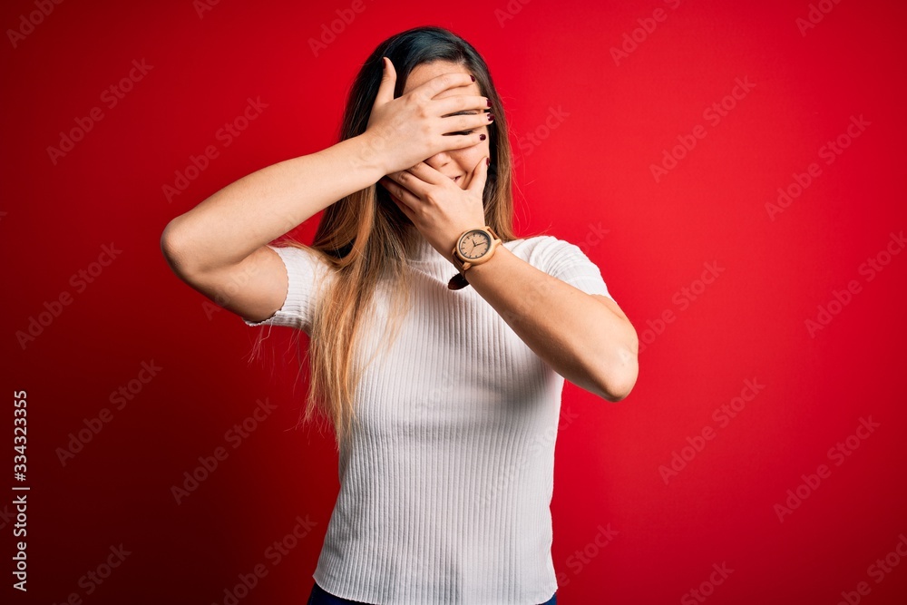 Beautiful blonde woman with blue eyes wearing casual white t-shirt over red background Covering eyes and mouth with hands, surprised and shocked. Hiding emotion Beautiful blonde woman with blue eyes wearing casual white t-shirt over red background Covering eyes and mouth with hands, surprised and shocked. Hiding emotion