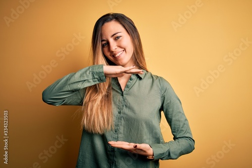 Young beautiful blonde woman with blue eyes wearing green shirt over yellow background gesturing with hands showing big and large size sign, measure symbol. Smiling looking at the camera. Measuring