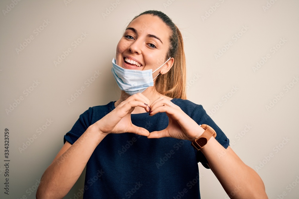 Beautiful blonde woman with blue eyes wearing medical mask over white background smiling in love showing heart symbol and shape with hands. Romantic concept. Beautiful blonde woman with blue eyes wearing medical mask over white background smiling in love showing heart symbol and shape with hands. Romantic concept.