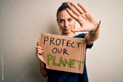 Young beautiful blonde woman with blue eyes asking for protect planet holding banner with open hand doing stop sign with serious and confident expression, defense gesture © Krakenimages.com