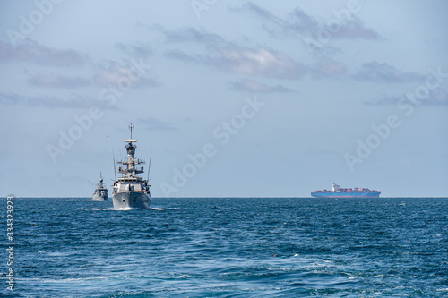 Two corvette warships sail together in the sea with container ship on the horizon