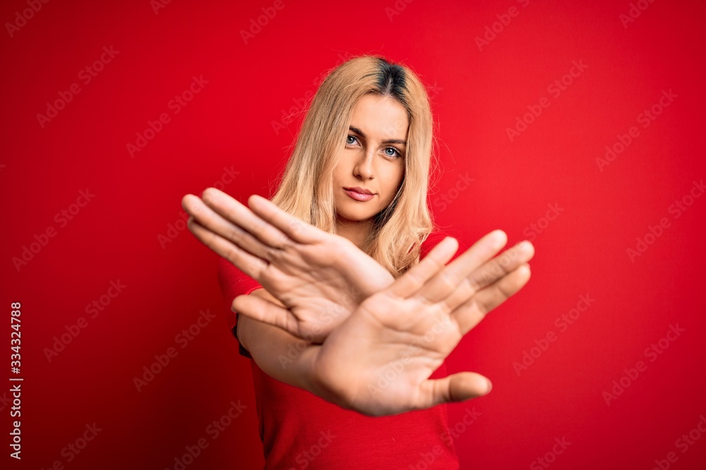 Young beautiful blonde woman wearing casual t-shirt standing over isolated red background Rejection expression crossing arms and palms doing negative sign, angry face Young beautiful blonde woman wearing casual t-shirt standing over isolated red background Rejection expression crossing arms and palms doing negative sign, angry face
