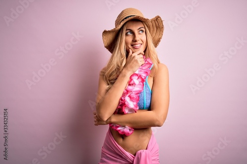 Young beautiful blonde woman on vacation wearing bikini and hat with hawaiian lei flowers with hand on chin thinking about question, pensive expression. Smiling with thoughtful face. Doubt concept.