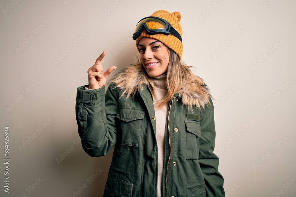 Young brunette skier woman wearing snow clothes and ski goggles over white background smiling and confident gesturing with hand doing small size sign with fingers looking and the camera. Measure Young brunette skier woman wearing snow clothes and ski goggles over white background smiling and confident gesturing with hand doing small size sign with fingers looking and the camera. Measure