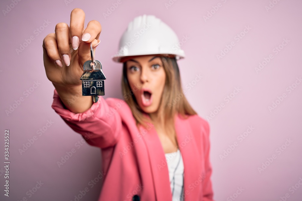Young beautiful brunette architect woman wearing safety helmet hoding key of house scared in shock with a surprise face, afraid and excited with fear expression