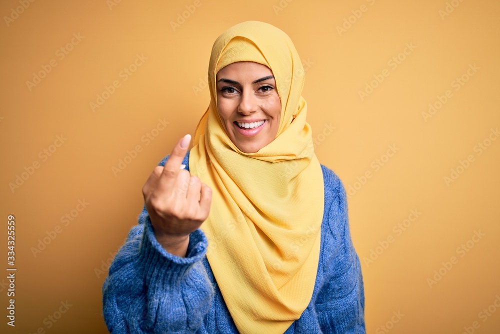 Young beautiful brunette muslim woman wearing arab hijab over isolated yellow background Beckoning come here gesture with hand inviting welcoming happy and smiling Young beautiful brunette muslim woman wearing arab hijab over isolated yellow background Beckoning come here gesture with hand inviting welcoming happy and smiling