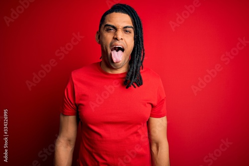 Young handsome african american afro man with dreadlocks wearing red casual t-shirt sticking tongue out happy with funny expression. Emotion concept.