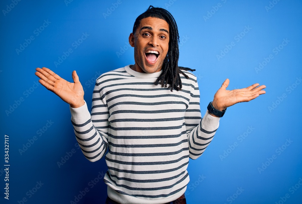 Naklejka premium Young handsome african american afro man with dreadlocks wearing casual striped sweater celebrating crazy and amazed for success with arms raised and open eyes screaming excited. Winner concept